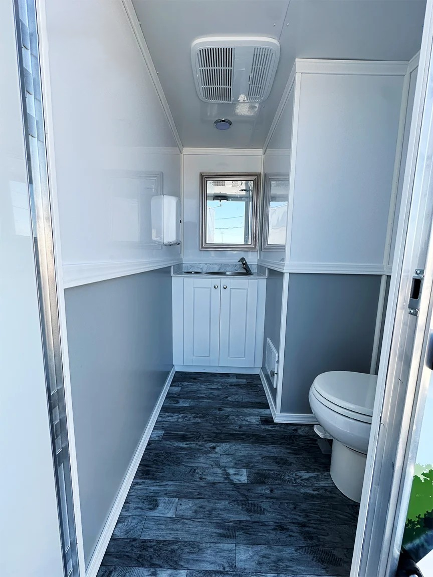 Interior view of 17-foot ADA restroom trailer featuring toilet, vanity sink cabinet, mirror, air vent, and wood-style flooring.