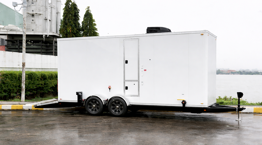 luxury restroom trailer parked near an industrial facility and waterfront area with dual axle wheels and rooftop air unit.
