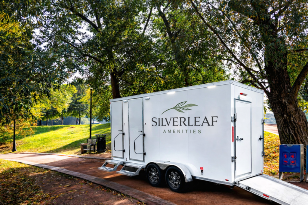 White luxury restroom trailer with dual entry doors, steps, and wheelchair ramp positioned along a park pathway surrounded by trees.
