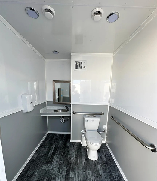 Interior of 17-foot ADA restroom trailer featuring accessible toilet with grab bars, stainless sink, mirror, ventilation fan, and wood-style flooring.