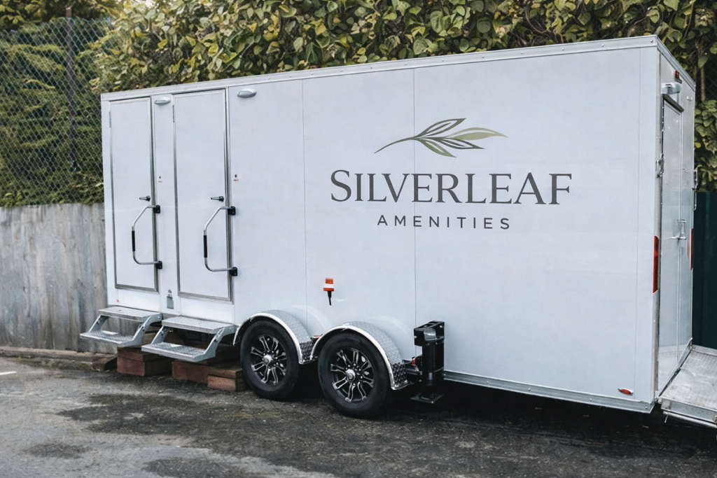 White luxury restroom trailer with dual entry doors and steps positioned in a parking area beside a wall and greenery.